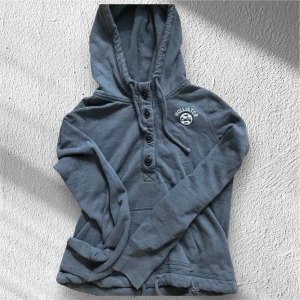 Grå hoodie från Hollister med knappar - Säljer en grå hoodie från Hollister med broderad logga på bröstet. Tröjan har huva med snörning, fyra svarta knappar framtill och långa ärmar. Mjuk bomullskänsla och fickor på sidorna. Perfekt för chill dagar och enkel att matcha.