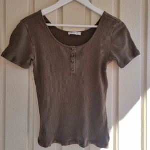 Ribbad brun topp - Toppen har rund halsringning och fyra knappar. Materialet är stretchigt med en enkel och stilren design. Det står storlek L men tycker den känns mindre på mig som har M oftast. 