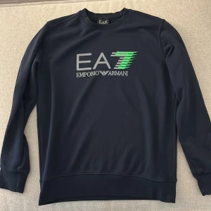 Mörkblå EA7 Emporio Armani sweatshirt - Snygg mörkblå sweatshirt från EA7 Emporio Armani med stort tryck på bröstet i grått och grönt. Tröjan har rund hals, långa ärmar och ribbade muddar. Perfekt för dig som gillar stilrena märkesplagg med sportig vibe.