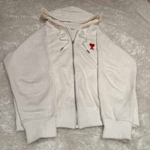 Vit Ami paris zip hoodie - Säljer en vit hoodie från Ami paris med dragkedja och huva. Tröjan har snörning vid halsen och ett broderat rött hjärta med bokstaven A på bröstet. Tillverkad i mjuk bomull. Tröjan är inprincip helt ny bara testad. Storlek L men sitter som M.