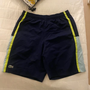 Blå Lacoste shorts med gula detaljer - Snygga marinblå shorts från Lacoste med ljusblå paneler på sidorna och gula ränder längs benen. Klassisk krokodil-logga framtill. Perfekta för sommaren och sportiga aktiviteter. Resår i midjan för skön passform.
