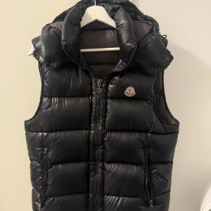 Svart dunväst från Moncler - Säljer en svart dunväst från Moncler med huva och dragkedja framtill. Västen har två fickor med dragkedja och Moncler-logga på bröstet. Perfekt för lager-på-lager och håller dig varm under kyliga dagar.