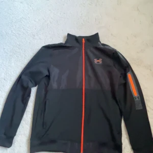 Svart vindjacka från Under Armour M - Svart vindjacka från Under Armour med orange dragkedja och logga på bröstet. Jackan har hög krage, lång ärm och en snygg detalj med orange dragkedja på ena ärmen. Perfekt för dig som gillar sportig stil.
