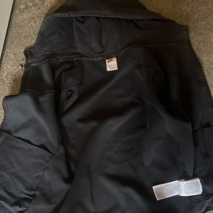 Svart Nike vindjacka med huva XL i barn storlek  - Svart vindjacka från Nike i storlek XL. Jackan har huva, dragkedja framtill och flera fickor, inklusive en med dragkedja på ärmen. Snyggt Nike-logga på bröstet och diskreta detaljer i svart. Perfekt för dig som gillar sportig och clean stil.