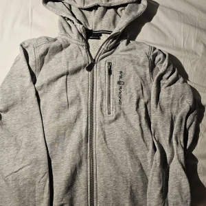 Grå hoodiejacka från Sail Racing - Grå hoodiejacka från Sail Racing med dragkedja framtill och bröstficka med dragkedja. Jackan har broderad logga på bröstet och är tillverkad i mjuk bomullsblandning. Perfekt för chill dagar och enkel att matcha med det mesta.