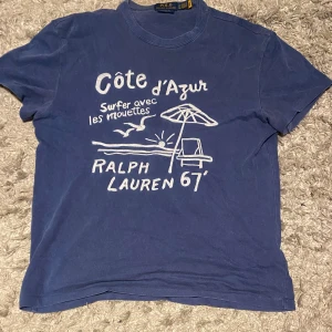 Blå t-shirt Polo Ralph Lauren - Blå t-shirt från Polo Ralph Lauren med vit Côte d'Azur-print och strandmotiv framtill. Klassisk rund halsringning och korta ärmar. Skön bomullskvalitet och relaxed vibe, perfekt för sommardagar. Otroligt bra skick, nypris ligger på runt 2000! Hör av er om ni har några frågor!