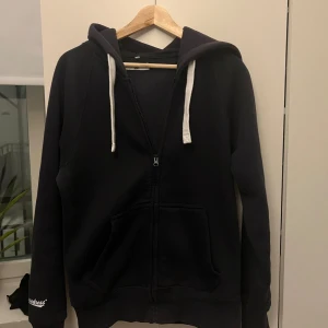 Mörkblå hoodie med dragkedja och huva - Mörkblå hoodie med vit snörning i huvan och dragkedja framtill. Tröjan har känguruficka och långa ärmar. Perfekt för chill dagar eller när du vill ha en enkel och stilren look.