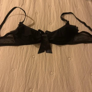 Svart  bh med volang och rosett - Supersöt svart bh från Axami 70 A med volangdetaljer i mesh och en stor glansig rosett framtill. Axelbandslös design med justerbara band som kan tas av. Perfekt för att ge en extra touch till din outfit.