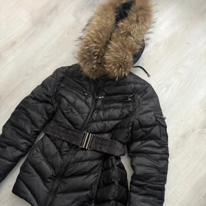 Svart pufferjacka med fuskpäls från hollies - Kan skicka fler bilder hur den sitter på privat.Svart pufferjacka från Jollies med stor huva kantad av brun fuskpäls. Jackan har markerad midja med brett bälte, dragkedjor framtill och på ärmen samt quiltad design. Perfekt för kalla dagar när du vill vara både varm och snygg.