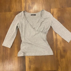 Grå omlottopp med trekvartsärm - Snygg grå omlottopp från Vero Moda med trekvartsärm och v-ringning. Topp i mjuk bomullsmix som sitter skönt och har en stilren look. Perfekt att matcha med jeans eller kjol för en enkel men trendig outfit.