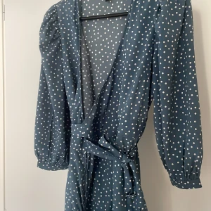 Blå prickig omlottklänning med volang - Snygg lång omlottklänning från Vero Moda i blått med vita prickar. Klänningen har puffiga långärmar, knytband i midjan och volangkant nedtill. V-ringad och med lätt, flowy passform som ger en chill vibe. Perfekt för dig som gillar retro och trendiga detaljer.