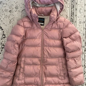 Rosa pufferjacka från Lassie - Säljer en fluffig rosa pufferjacka från Lassie med huva och dragkedja. Jackan har två fickor framtill och är perfekt för kalla dagar. Den är lätt vadderad och har en snygg, enkel design som håller dig varm och bekväm.