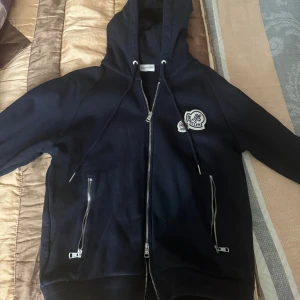 Mörkblå Moncler hoodie med dragkedja - Säljer en mörkblå hoodie från Moncler med huva och snygga silverfärgade dragkedjor framtill och vid fickorna. Tröjan har Moncler-logga på bröstet och justerbara snören vid huvan. Perfekt för dig som gillar exklusiva streetwear-vibbar. Använt vid två tillfällen och sedan bara legat i garderoben