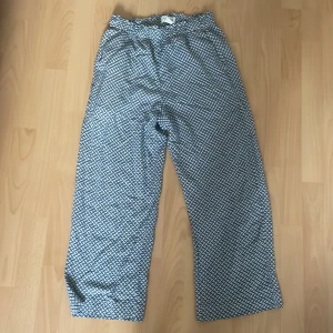 Mönstrade byxor  - Snygga byxor från Holly & Whyte by Lindex i storlek 36. De är vita med mörkblått blommigt mönster, har hög midja med resår och raka kortare ben. Materialet är mjukt och luftigt, perfekt för en chill och trendig look.