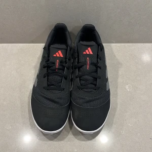 Adidas Predator svart inneskor - Säljer ett par Adidas Predator inneskor i svart med gråa ränder och röda detaljer. Skorna har snörning, platt sula och rund tå. Ovandelen är i syntetmaterial och mesh för bra ventilation. Perfekt för inomhusspel och snygg stil på planen.