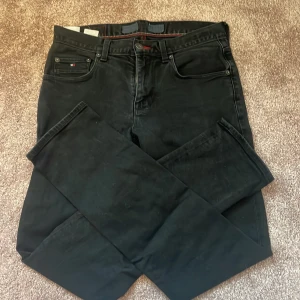 Svarta straight fit jeans Tommy Hilfiger - Svarta jeans från Tommy Hilfiger i straight fit med stretch. Klassisk femficksmodell med diskret logga på myntfickan och snygga röda sömmar vid linningen. Perfekt passform och stilren look för dig som gillar enkel och clean stil.