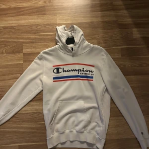 Vit Champion hoodie - Fräsch vit hoodie från Champion med tryck i blått och rött framtill. Bekväm och stilren modell med känguruficka och justerbar huva. I mycket fint skick, endast använd ett fåtal gånger. Inga fläckar eller slitage.