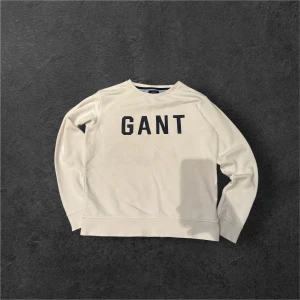 Vit sweatshirt från GANT med tryck - Vit sweatshirt från GANT med svart tryck över bröstet. Tröjan har rund hals, långa ärmar och ribbade muddar vid ärmslut och nederkant. Tillverkad i mjuk bomull, perfekt för chill dagar. Klassisk och enkel design med snygg logga framtill.