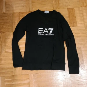 Svart långärmad tröja EA7 Armani - Svart långärmad tröja från EA7 Emporio Armani med vit logga framtill och tryck på ryggen. Klassisk rund halsringning och mjukt material som känns skönt mot huden. Perfekt för dig som gillar stilrena och snygga plagg med premiumkänsla.