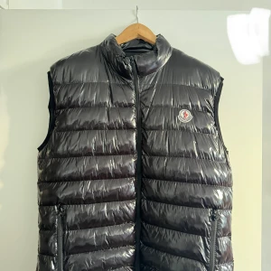 Svart dunväst från Moncler - Snygg svart dunväst från Moncler med glansig finish och horisontella quiltade paneler. Västen har hög krage, dragkedja framtill och två fickor med dragkedja. Moncler-logga på bröstet ger en exklusiv vibe. Perfekt för lager-på-lager under kyliga dagar.