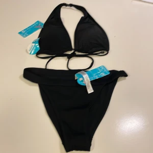 Svart bikini från New Yorker  med ribbad detalj - Snygg svart bikini från New Yorker  med klassisk trekants-bh och knyt i ryggen. Underdelen har ribbad struktur och bredare kant upptill för extra stilpoäng. Perfekt för dig som gillar enkel och stilren look på stranden. Materialet är mjukt och följsamt syntet.