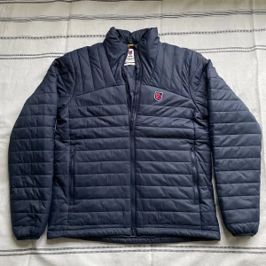 Mörkblå Dunjacka Fjällräven M - Säljer en helt oanvänd dunjacka från Fjällräven. Storlek M, orginalförpakning eller kvitto finns ej kvar. Fick den som present, men kom aldrig till användning.