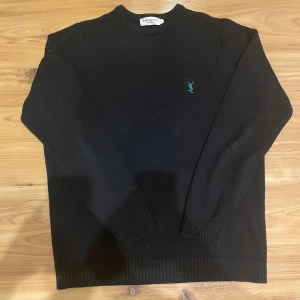 Svart stickad tröja från Yves Saint Laurent - Svart stickad tröja från Yves Saint Laurent med rund hals och ribbade muddar. På bröstet finns en broderad YSL-logga i turkos. Tröjan har en klassisk och clean look, perfekt för dig som gillar stilrena designer och exklusiva märken. Skicket är mycket bra och har inga defekter! Om ni har några frågor så är det bara att höra av er😁