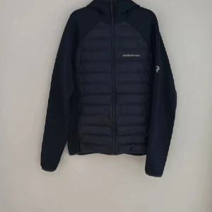 Peak Performance Hybridjacka - Svart hybridjacka från Peak Performance i storlek M. Jackan har quiltad framsida och rygg, huva och dragkedja framtill. Ärmarna är i mjukare material och en blå detaljsöm syns på ena ärmen. Perfekt för kyliga dagar.