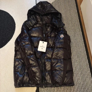 Svart Moncler dunjacka med huva M - Svart glansig dunjacka från Moncler i storlek M. Jackan har huva, dragkedja framtill, fickor med dragkedja och en extra ficka med knapp på ärmen. Klassisk Moncler-logga på ärmen och quiltad design. Perfekt för kalla dagar.
