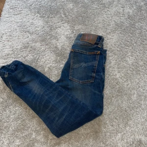 W26 L30Blå jeans med slitningar från Nudie - Snygga blå jeans från Nudie med slitna detaljer och klassisk femficksdesign. Jeansen har orange kontrastsömmar, läderpatch bak och en rak passform. Materialet är robust denim i bomull och de har coola slitningar på ben och ficka.