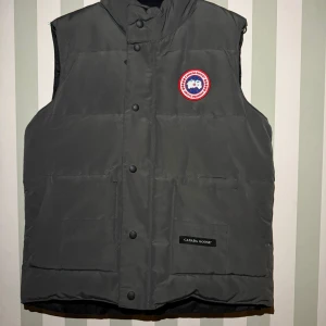Mörkgrå dunväst från Canada Goose - Snygg mörkgrå dunväst från Canada Goose med klassisk logotyp på bröstet. Västen har hög krage, tryckknappar framtill och två stora fickor. Perfekt för kalla dagar och har en stilren, quiltad design. Materialet är slitstark polyester och dunfyllning.