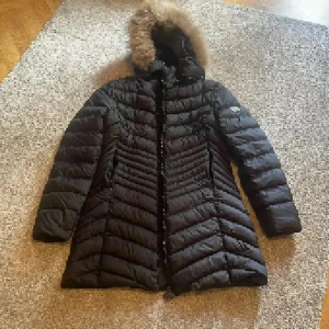 Svart dunjacka från Moncler med fuskpäls - Säljer en svart lång dunjacka från Moncler med snyggt quiltat mönster, huva med fuskpäls och tvåvägsdragkedja med logga. Jackan har lång ärm och är perfekt för kalla vinterdagar. Klassisk och stilren design som håller dig varm hela säsongen. Nfc funkar😊