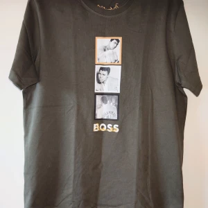 Khaki BOSS x Muhammad Ali t-shirt 3XL, oanvänd  - Khaki t-shirt från BOSS x Muhammad Ali med tryck av tre svartvita foton och texten 'BOSS' i vitt och guld på framsidan. Klassisk rund hals och korta ärmar. Tillverkad i mjuk bomull för en skön känsla. Aldrig använd, endast testat. Han tvätta den en gång. 