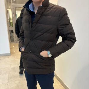 Mörkbrun quiltad kavajjacka - Snygg mörkbrun pufferjacka med quiltad design och raka linjer. Jackan har ståkrage, två sidofickor och knäppning med knappar framtill. Perfekt för dig som vill ha en stilren look under kyliga dagar. Passar dig som gillar klassisk och enkel stil. En biligare variant istället för att köpa en herno 