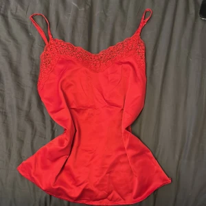 Röd spetslinne från Tezenis - Supersnygg röd spetslinne från Tezenis med tunna axelband och vacker spetsdetalj upptill. Linnet har en slits på sidan och är gjord i ett glansigt, mjukt material som känns lyxigt. Perfekt för dig som vill sticka ut med en feminin och trendig look.