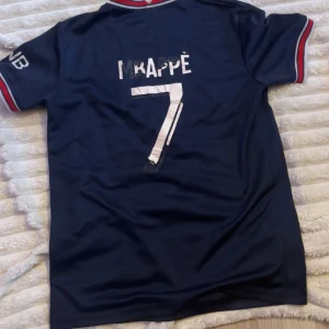 PSG Mbappé matchtröja blå - Snygg PSG fotbollströja i mörkblått med röda och vita detaljer på krage och ärmslut. Tryck med Mbappé och nummer 7 på ryggen. Officiell klubbmärke på bröstet och sponsortryck framtill. Tillverkad i lätt och ventilerande material, perfekt för fotboll eller support. Tröjan är väldigt sliten, därför är priset också lågt. Pris kan diskuteras!