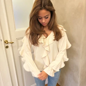 Krämvit blus med volanger - Säljer en krämvit blus med v-ringning och stora volangdetaljer längs halsringningen och ärmarna. Blusen har långärmade, lätt puffade ärmar och är tillverkad i ett mjukt, lätt material som ger ett flowy intryck. Perfekt att styla med jeans eller kjol.