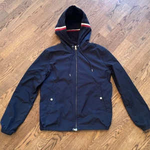 Mörkblå vindjacka från Moncler - Snygg mörkblå vindjacka från Moncler med huva och dragkedja. Jackan har Moncler-logga på ärmen, två fickor med tryckknappar och elastiska muddar. Huvan har en röd och vit randig detalj som ger en sportig vibe. Perfekt för dig som gillar stilrena och exklusiva plagg. Stl 1 motsvarar S
