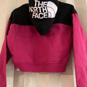 Svart och cerise hoodie från The North Face - Cool hoodie från The North Face i svart och cerise med stor logga på huvan och mindre logga på bröstet. Tröjan har croppad passform, långa ärmar och ribbad nederkant. Perfekt för dig som gillar streetstyle och vill sticka ut.