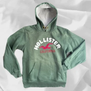 Grön hoodie från Hollister - Snygg grön hoodie från Hollister med vit och rosa brodyr på bröstet. Tröjan har huva med snörhål, magficka och ribbade muddar. Perfekt för chill dagar och streetstyle.