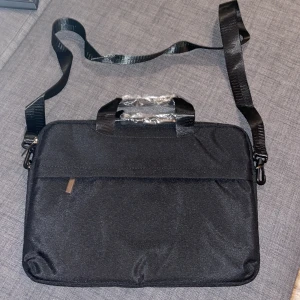 Svart datorväska med axelrem - En stilren svart datorväska med justerbar axelrem och två handtag. Väskan har en enkel design med dragkedja och en praktisk ytterficka framtill. Perfekt för att bära laptop och tillbehör. Materialet är slitstark polyester. Helt ny jag säljer den då jag råkat beställa i fel storlek till min dator 