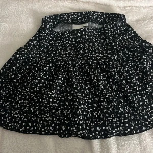 Svart blommig kortkjol från Gina Tricot - Supersöt svart kortkjol från Gina Tricot med vitt blommigt mönster🌸🌸  Storlek 146/152, men sitter perfekt för mig som egentligen har storlek S. Den är stretchig vilket gör att den sitter väldigt fint och perfekt! Kjolen har volangdetaljer och en bekväm, elastisk midja. Perfekt att matcha med en enkel topp eller hoodie. Materialet är mjukt och lätt, vilket gör den skön att bära under varma dagar. Typ aldrig använd❣️💕 Väldigt bra skick!