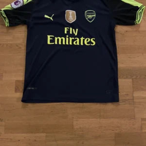 Arsenal fotbollströja Puma marinblå - Snygg Arsenal fotbollströja från Puma i marinblått med neongula detaljer och tryck. Kortärmad modell med klubbmärke, FIFA 2018 badge och Fly Emirates sponsor på bröstet. Tillverkad i lätt och ventilerande polyester, perfekt för match eller träning.