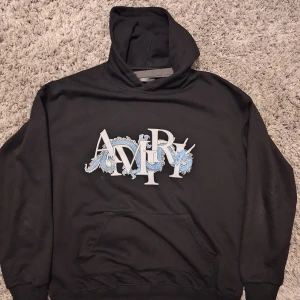 Svart hoodie från Amiri med draktryck - Svart hoodie från Amiri med stor logga och blå drakmotiv på bröstet. Klassisk känguruficka framtill och huva. Tröjan har långa ärmar och en relaxed passform, perfekt för en streetwear-look.