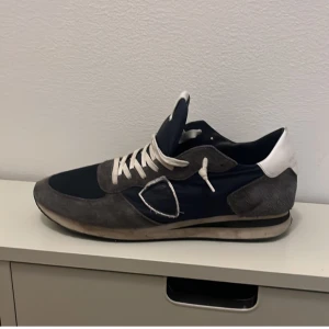 Philippe Model trainers - Feta trainers från Philippe Model i marinblått och grått med vita detaljer. Skorna har klassisk snörning, rund tå och platt sula. Ovandelen är i en mix av mocka och textil, med vit häl och logga på sidan. Perfekta för en stilren och avslappnad look.