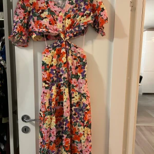 Blommig långklänning från Lindex - Färgstark långklänning från Lindex med blommigt mönster i rosa, lila, röd, orange och gul. Klänningen har omlott-detalj i midjan, korta puffärmar och v-ringad hals. Materialet är lätt och luftigt, perfekt för varma dagar. Snygg cut-out i midjan för extra edge.