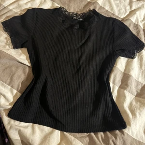 Svart ribbad topp med spets från Zara - Supersnygg svart ribbad topp från Zara med korta ärmar och fina spetsdetaljer vid halsringning och ärmslut. Toppen har en figurnära passform. Passar bra ändå om man är XS. 