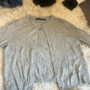 Grå kofta från 157 XS - Snygg grå kofta från 157 i storlek XS. Den har rund hals, långa ärmar och knappar framtill. Materialet är mjukt och stickat, med ribbade muddar vid ärmslut och nederkant. Perfekt att slänga över en t-shirt när det blir lite kyligt.