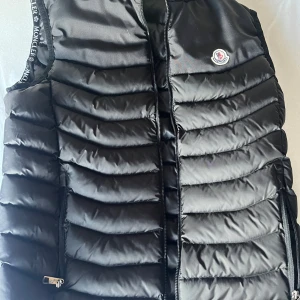 Svart dunväst från Moncler M - Svart dunväst från Moncler med quiltad design och dragkedja framtill. Västen har broderad Moncler-logga på bröstet, stor brodyr på ryggen och Moncler-detaljer längs ärmöppningarna. Perfekt för lager-på-lager och snygg streetstil.