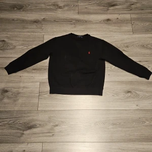 Svart tröja från Polo Ralph Lauren - Svart långärmad tröja från Polo Ralph Lauren med klassisk rund halsringning och röd broderad logga på bröstet. Tröjan är i mjuk bomull och har ribbade muddar vid ärmslut och nederkant. Perfekt för en clean och stilren look.